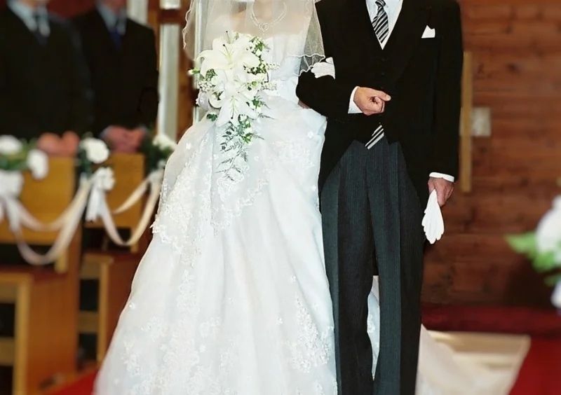 白いウェディングドレスとベールを着た花嫁が白いユリのブーケを持ち、黒いモーニングコートを着た新郎と腕を組んで教会のバージンロードを歩いている