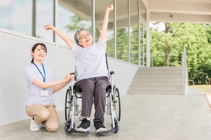 車椅子に座り両腕を上げてストレッチする眼鏡をかけた高齢女性と、青いストラップの介護スタッフが笑顔でサポートしている明るい屋内施設